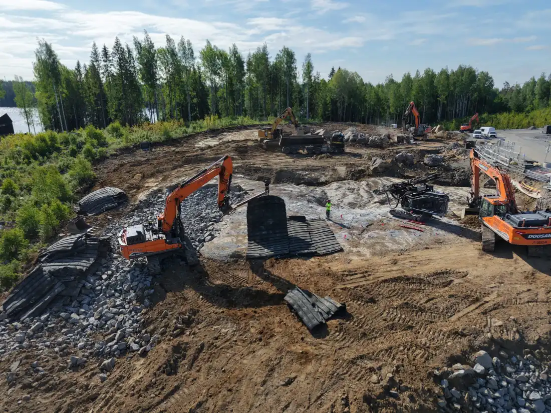 Ilmakuva louhintatyömaasta, jossa kaivinkoneet ja porauslaite työskentelevät järven rannalla